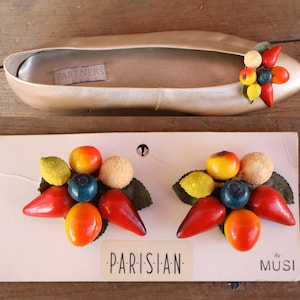 May include: A gold-toned flat shoe with a label reading PARTNERS inside, featuring a cluster of miniature fruit embellishments on the toe. Below, two matching fruit-themed shoe clips are attached to a card labeled PARISIAN by MUSI.