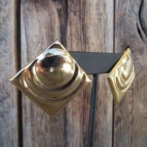 Puede incluir: Un par de pendientes de tono dorado con un diseño geométrico. Los pendientes son de forma cuadrada con un centro redondeado y una superficie texturizada.