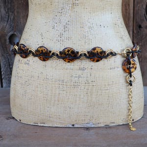 May include: A vintage belt with a tortoiseshell design. The belt features circular tortoiseshell accents connected by gold-tone and black chain links. A gold chain dangles from the belt's end, adding a decorative touch. The belt is displayed on a mannequin.