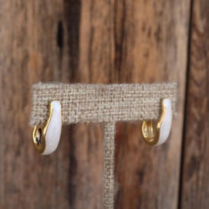 May include: A pair of gold hoop earrings with white enamel. The earrings are displayed on a burlap earring stand.