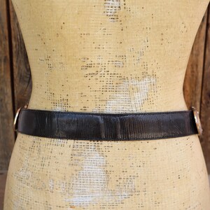 May include: A black belt with a textured surface and gold-colored buckle. The belt is wrapped around a mannequin. The background is a wooden wall.