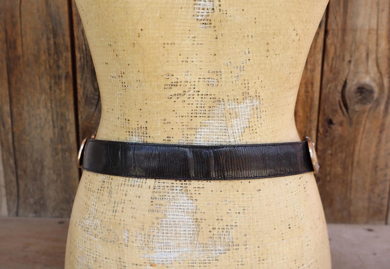 May include: A black belt with a textured surface and gold-colored buckle. The belt is wrapped around a mannequin. The background is a wooden wall.