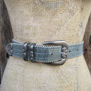 May include: A gray leather belt with a silver buckle. The buckle has a decorative design with swirls.