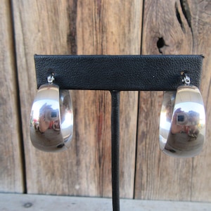 May include: A pair of polished silver hoop earrings. The earrings are displayed on a black stand against a wooden background. The hoops are thick and have a shiny, reflective surface, capturing the surrounding light and colors.