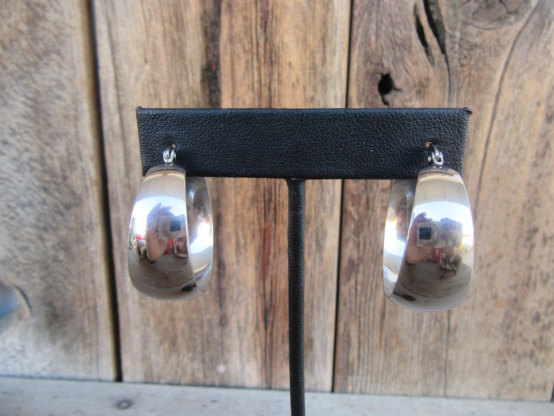May include: A pair of polished silver hoop earrings. The earrings are displayed on a black stand against a wooden background. The hoops are thick and have a shiny, reflective surface, capturing the surrounding light and colors.