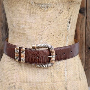 May include: Brown leather belt with a textured pattern, featuring a silver and gold-toned buckle. The belt is displayed on a mannequin against a rustic wooden background. The belt has a classic design.