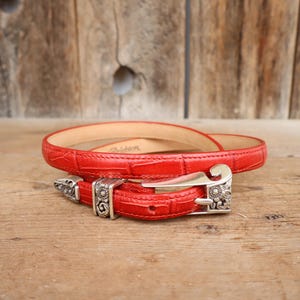 Puede incluir: Un cintur&oacute;n de cuero rojo con un patr&oacute;n texturizado similar al de un cocodrilo. El cintur&oacute;n presenta una hebilla plateada con detalles ornamentados. El cintur&oacute;n est&aacute; enrollado sobre una superficie de madera.