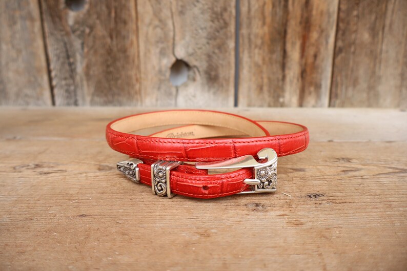 Puede incluir: Un cintur&oacute;n de cuero rojo con un patr&oacute;n texturizado similar al de un cocodrilo. El cintur&oacute;n presenta una hebilla plateada con detalles ornamentados. El cintur&oacute;n est&aacute; enrollado sobre una superficie de madera.