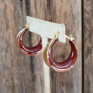 May include: Gold-tone hoop earrings with a glossy, deep burgundy enamel interior. The earrings are displayed on a neutral stand, highlighting their curved, crescent shape. The earrings have a classic, elegant design, suitable for various occasions.