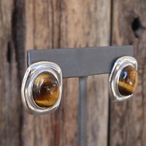 May include: A pair of silver earrings with tiger's eye stones. The earrings are round and have a smooth, polished finish.