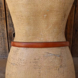 May include: A brown leather belt is wrapped around a mannequin. The belt is a medium brown color and appears to be made of smooth leather. The mannequin is a light tan color and has a vintage appearance. The background is a wooden wall.
