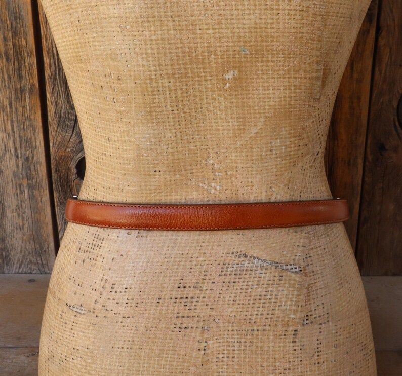 May include: A brown leather belt is wrapped around a mannequin. The belt is a medium brown color and appears to be made of smooth leather. The mannequin is a light tan color and has a vintage appearance. The background is a wooden wall.