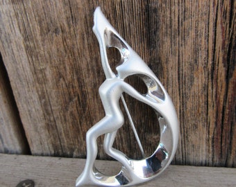 Puede incluir: Broche plateado con una figura humana estilizada en una pose elegante. La figura está integrada en un diseño en forma de media luna con espacios abiertos, creando una estética moderna y artística. El broche tiene un cierre de alfiler en la parte posterior.