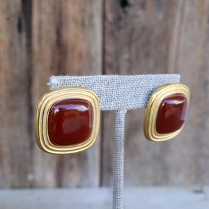 May include: A pair of square-shaped earrings with a gold-tone border and a rich, reddish-brown center. The earrings are displayed on a gray earring stand against a blurred wooden background. The design is classic and elegant.