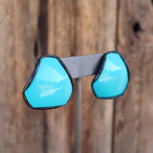 Puede incluir: Un par de pendientes de clip azul turquesa con marcos de metal negro. Los pendientes tienen forma de pentágonos irregulares.