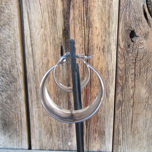May include: A pair of silver hoop earrings. The earrings are displayed on a black stand against a backdrop of weathered wooden planks. The hoops have a smooth, polished finish and a classic, rounded shape.
