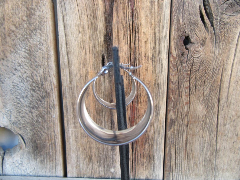 May include: A pair of silver hoop earrings. The earrings are displayed on a black stand against a backdrop of weathered wooden planks. The hoops have a smooth, polished finish and a classic, rounded shape.