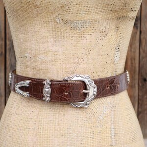 Op de afbeelding: Bruine leren riem met een zilverkleurige gesp en accenten. De riem heeft een krokodillenleertextuur en wordt op een mannequin getoond. De gesp is sierlijk en gedetailleerd.