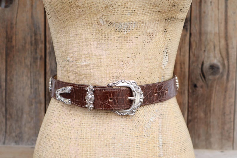 Op de afbeelding: Bruine leren riem met een zilverkleurige gesp en accenten. De riem heeft een krokodillenleertextuur en wordt op een mannequin getoond. De gesp is sierlijk en gedetailleerd.
