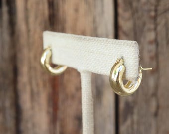 May include: A pair of gold-colored hoop earrings. The earrings are thick and rounded, with a polished finish. They are displayed on a white fabric stand against a blurred wooden background.