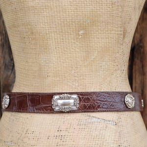 Op de afbeelding: Een bruine leren riem met een krokodillenstructuur. De riem heeft een zilveren rechthoekige gesp met sierlijke details en zilveren cirkelvormige accenten. De riem wordt op een mannequin getoond.