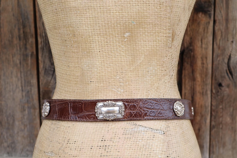 Op de afbeelding: Een bruine leren riem met een krokodillenstructuur. De riem heeft een zilveren rechthoekige gesp met sierlijke details en zilveren cirkelvormige accenten. De riem wordt op een mannequin getoond.
