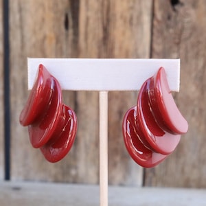 Puede incluir: Un par de pendientes rojos vintage. Cada pendiente presenta un diseño curvo y en capas, con un acabado brillante. Los pendientes se exhiben en un soporte blanco, sobre un fondo de madera. Un accesorio llamativo.