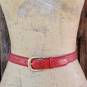 May include: A red belt with a textured snakeskin pattern and a gold-toned metal buckle. The belt features a matching red leather loop to secure the end of the strap and multiple adjustment holes along the length of the band.