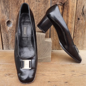 Könnte beinhalten: Ein Paar schwarze Lederpumps mit eckiger Spitze und silberfarbener Schnalle. Die Schuhe haben einen Blockabsatz. Der Markenname "BALLY" ist im Inneren eines Schuhs sichtbar. Die Schuhe stehen auf einer Holzoberfläche.