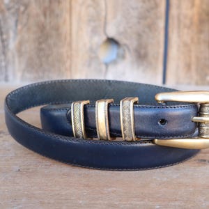 May include: A navy blue leather belt with a gold-colored buckle and three gold-colored belt loops. The belt is coiled on a wooden surface. The buckle has a textured design.