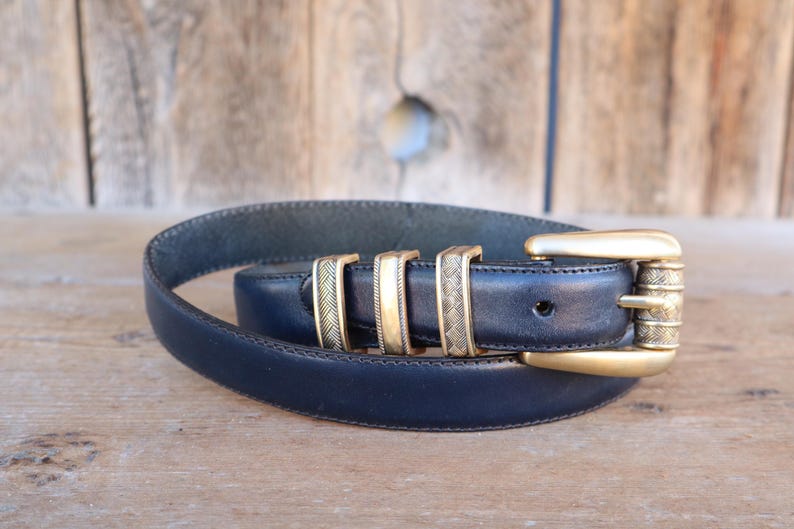 May include: A navy blue leather belt with a gold-colored buckle and three gold-colored belt loops. The belt is coiled on a wooden surface. The buckle has a textured design.