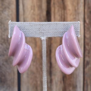 May include: A pair of light purple, sculptural earrings. Each earring has a curved, layered design with a glossy finish. The earrings are displayed on a neutral-colored, textured surface against a wooden background.