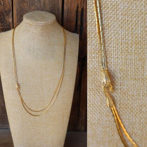 May include: A gold-toned necklace with a delicate chain design. The necklace is displayed on a beige mannequin. The necklace has a unique clasp and a layered design, adding a touch of elegance to the jewelry.