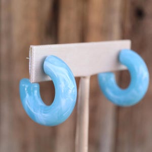 May include: A pair of light blue hoop earrings with a marbled white pattern. The earrings are displayed on a neutral-colored earring holder against a blurred wooden background. The earrings have a rounded, chunky design.