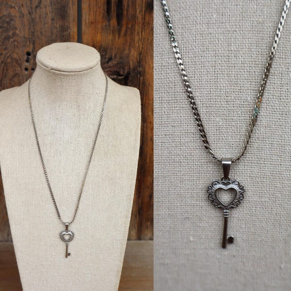May include: Silver-toned necklace featuring a key pendant with a heart-shaped design. The key has an ornate, openwork heart at the top and a simple key shape below. The necklace is displayed on a mannequin and against a wooden background.