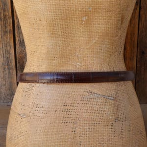 May include: A brown leather belt with a crocodile skin texture is wrapped around a mannequin. The belt is a dark shade of brown and appears to be made of high-quality leather. The belt is a classic accessory.