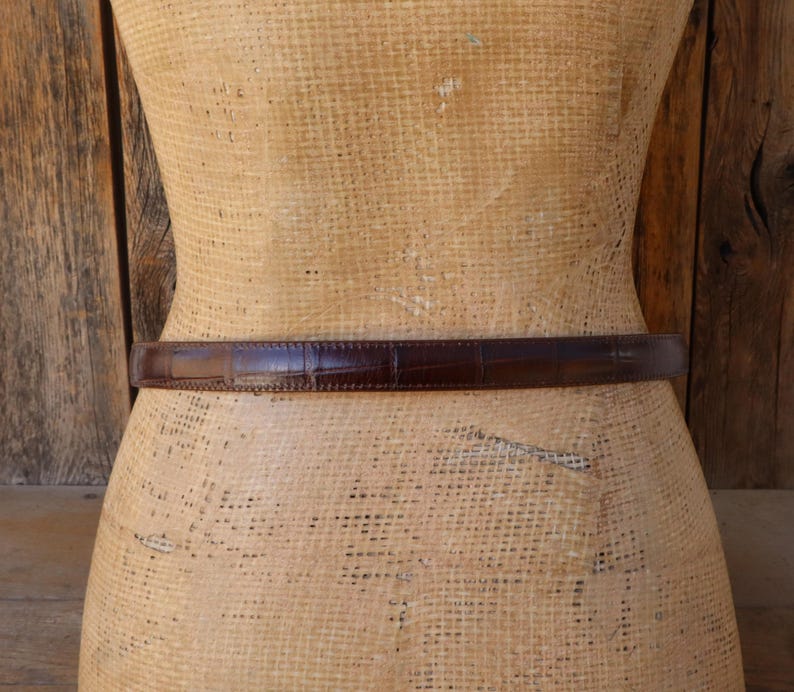 May include: A brown leather belt with a crocodile skin texture is wrapped around a mannequin. The belt is a dark shade of brown and appears to be made of high-quality leather. The belt is a classic accessory.