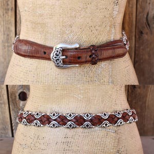 May include: Two brown leather belts with silver-tone metal accents. The top belt has a textured pattern and a decorative buckle. The bottom belt features a woven design with ornate silver embellishments. Both belts are displayed on a mannequin.
