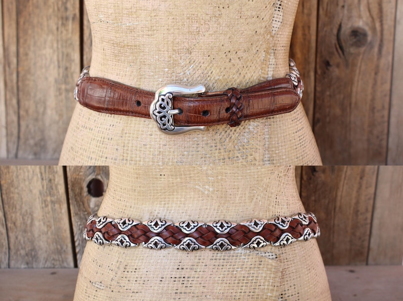 May include: Two brown leather belts with silver-tone metal accents. The top belt has a textured pattern and a decorative buckle. The bottom belt features a woven design with ornate silver embellishments. Both belts are displayed on a mannequin.