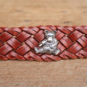 May include: A brown leather belt with a woven pattern. A silver bear charm is attached to the belt. The bear is depicted sitting and holding a small item. The belt is shown on a wooden surface.