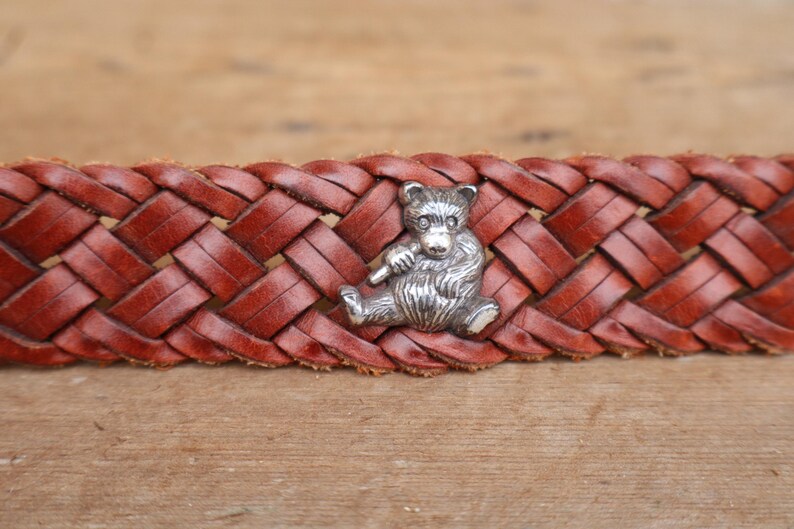 May include: A brown leather belt with a woven pattern. A silver bear charm is attached to the belt. The bear is depicted sitting and holding a small item. The belt is shown on a wooden surface.