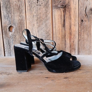 May include: A pair of black velvet heeled sandals with a peep-toe design. The sandals have a thick block heel and delicate straps with a gold-colored buckle. The background is a rustic wooden surface.