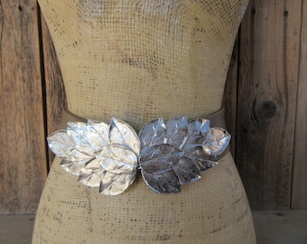 May include: A silver-colored leaf-shaped belt buckle is the focal point of this image. The buckle is attached to a neutral-toned belt. The buckle's design features multiple overlapping leaves. The background is a wooden surface.