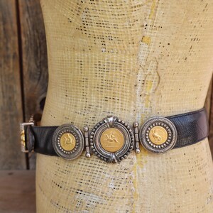 May include: Black leather belt with silver and gold-toned decorative accents. The belt features three circular medallions with gold-colored coin-like centers and a central oval medallion. The belt is displayed on a mannequin.
