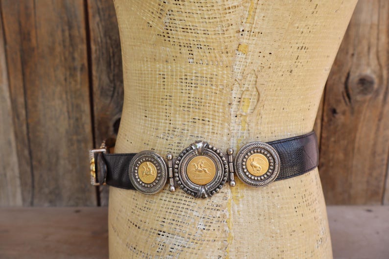 May include: Black leather belt with silver and gold-toned decorative accents. The belt features three circular medallions with gold-colored coin-like centers and a central oval medallion. The belt is displayed on a mannequin.