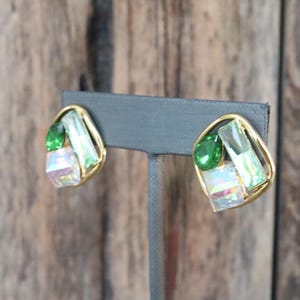 K&ouml;nnte beinhalten: Ein Paar goldfarbene Ohrringe mit gr&uuml;nen und schillernden Glassteinen. Die Ohrringe befinden sich auf einem schwarzen Pr&auml;sentationsst&auml;nder.
