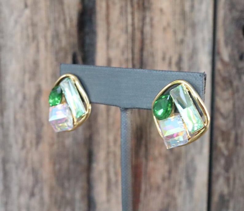 K&ouml;nnte beinhalten: Ein Paar goldfarbene Ohrringe mit gr&uuml;nen und schillernden Glassteinen. Die Ohrringe befinden sich auf einem schwarzen Pr&auml;sentationsst&auml;nder.