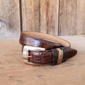 May include: A brown leather belt with a crocodile pattern. The belt has a gold-colored buckle with an ornate design and a woven leather loop. The belt is displayed on a wooden surface.