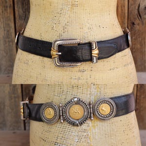 May include: Two black leather belts with decorative silver and gold buckles. The top belt has a large rectangular buckle with gold accents. The bottom belt features three circular medallions with gold-toned details and a central medallion with a horse design.