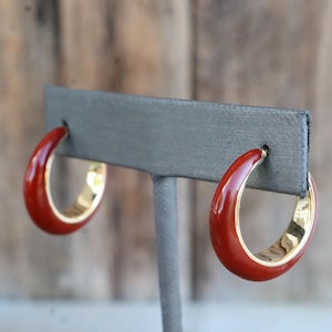 May include: A pair of red hoop earrings with a glossy finish. The earrings have a gold-colored interior and are displayed on a gray earring stand. The background is a blurred wooden surface.
